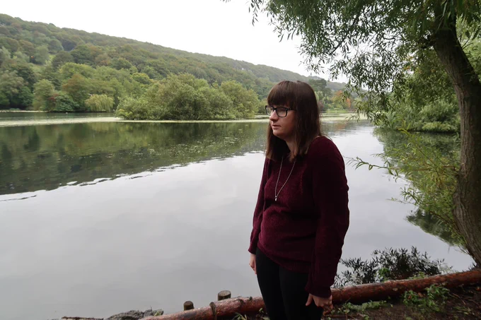 Holly stood under a willow tree looking to the right, there is a lake and trees behind her, she is wearing a burgundy jumper, black leggings and a silver necklace