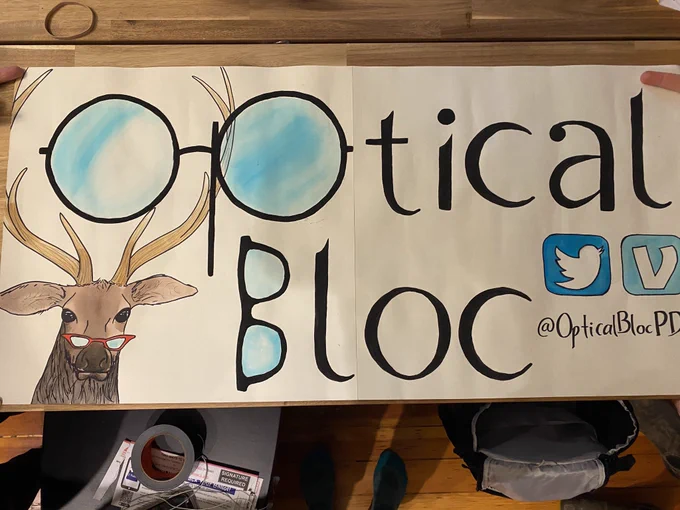 a poster sits on a table, it reads "Optical Bloc, @opticalblocPDX" and has images of the twitter and venmo icon above my handle. an elk with glasses looks out from the left corner. the "O" and "P" of Optical are drawn from a pair of glasses. the "B" in Bloc is also made from a pair of glasses. in the background, someone's backpack and a roll of duct tape sit on the ground.