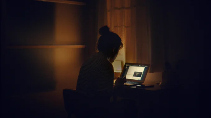 A woman sitting at her computer in the dark with a lamp on in the corner. 