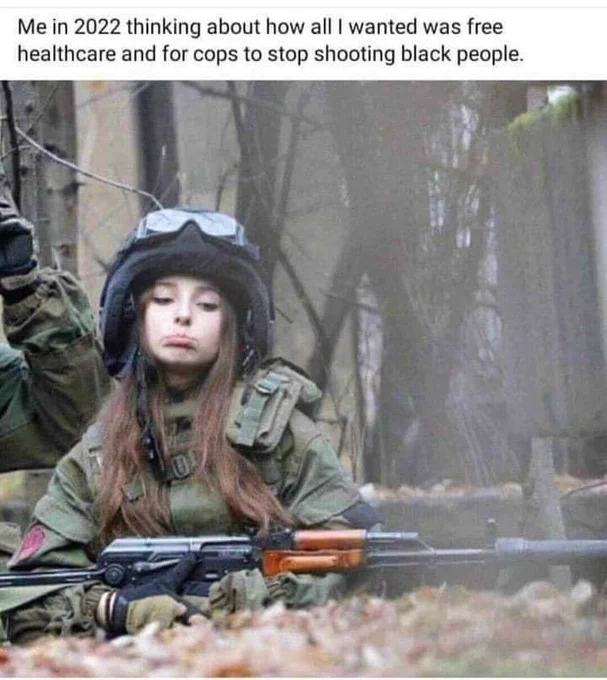 Image shows young woman in battle fatigues, including a helmet with goggles. She has long brown hair and is pouting. She is seen holding a long rifle with a silencer. She appears to be in a prone position ready to fire with a spotter looking down range. Text reads "me in 2022 thinking about how all I wanted was free healthcare and for cops to stop shooting Black people."
