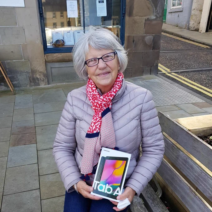 Photo of a Digital Buddies participant wearing a patterned red and pink scarf, smiling and sitting outside on a bench holding a tablet she received through the project.