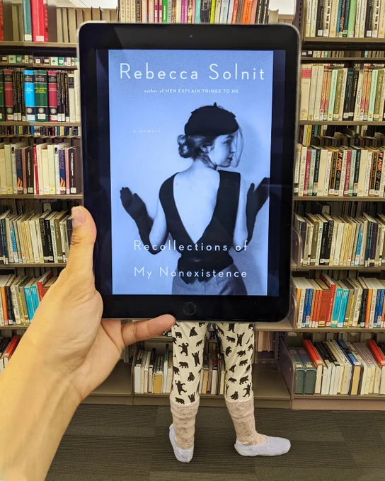 Recollections of My Non-Existence by Rebecca Solnit held in front of a person wearing pjs, fluffy socks and slippers.