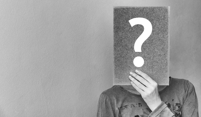 A grey image of a woman holding up a dark gray paper with a white question mark in front of  her face.