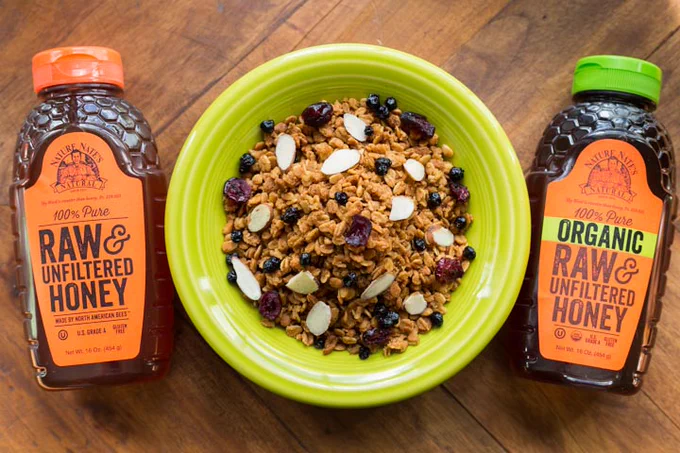 Bowl of honey granola next to two bottles of Nature Nate's honey on a wood table