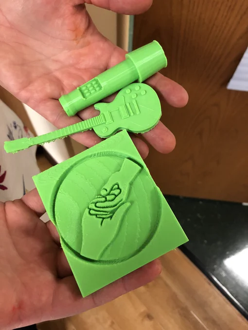 Hands outstretched holding 3 green 3D printed symbols. At the top is a microphone, in the middle is a guitar, at the bottom are hands holding each other.