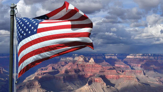 An American flag waves in the wind, with a view of a vast and colorful canyon in the distance.