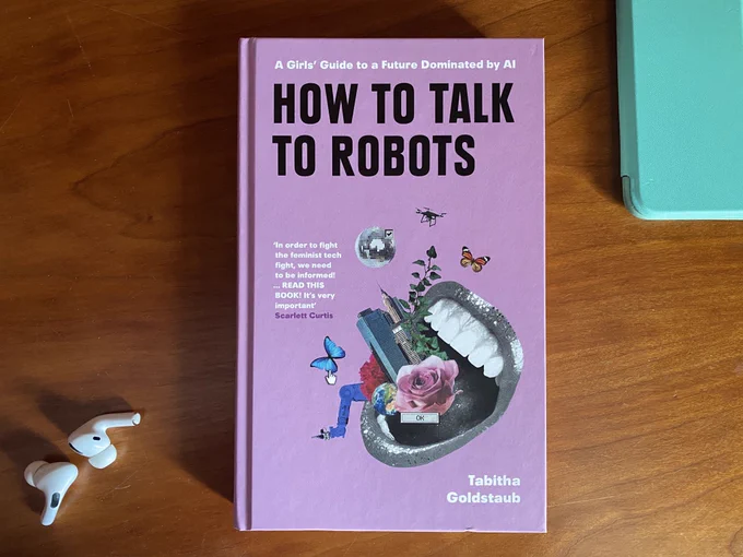 Picture of the book How To Talk To Robots by Tabitha Goldstaub on a wooden desk with Apple Airpords in the bottom left hand corner and the corner of a turquoise iPad cover peeking in the top right corner.