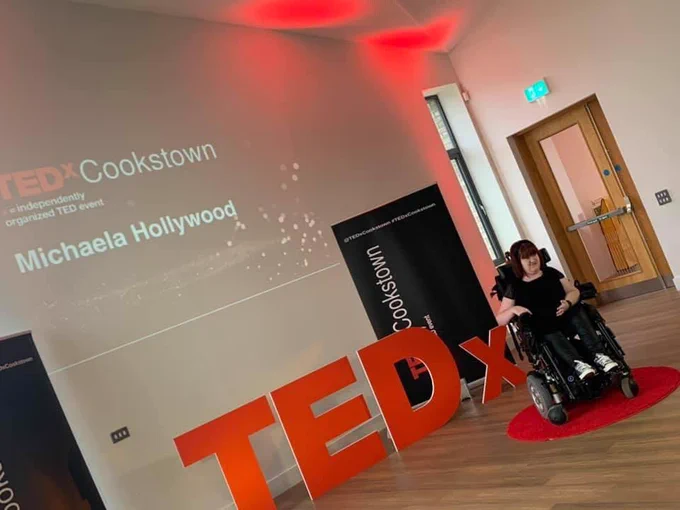 Michaela a white wheelchair user sits on a red dot with a red “TEDx” behind her. Her name is up behind her.