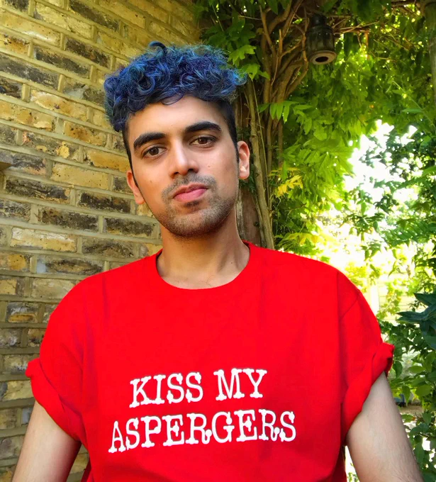 Image description: Vijay is sitting, head slightly tilted. He has brown skin, curly blue hair (medium length) and a bit of stubble on his face. He is wearing a red t shirt which has the words "KISS MY ASPERGERS" written on the centre of it, in white writing. 