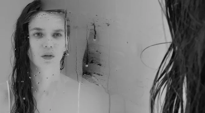 A black and white photo of a woman with dark, wet hair looking at her reflection in a partially steamy mirror.