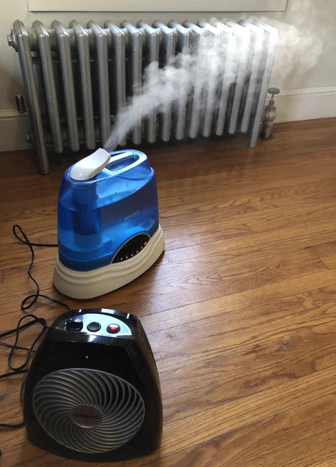 A space heater on a hardwood floor with a humidifier blowing a plume of water vapor out behind it. In the background is an old metal steam radiator.