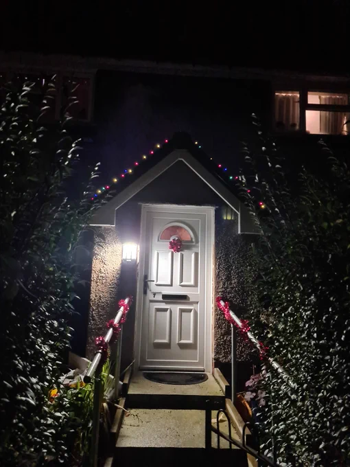 House front path with red tinsel on the bannisters of the front steps up to the front door which has a red bow on it. Coloured fairy light are draped over the gabled porch.