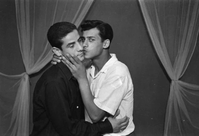 A black and white portrait of two men. One is kissing the other on the cheek.