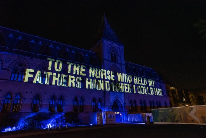 Illuminated message on the facade of the Oxford Museum of Natural History saying To the nurse who held my fathers hand when I could not in cream letters on blue background