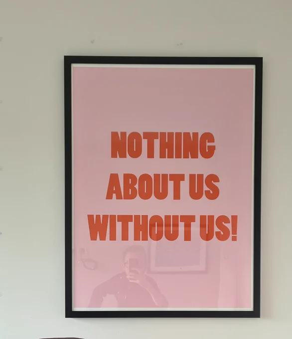Self portrait reflection. Sean taking a photo of a framed poster in his office. The poster reads Nothing About Us Without Us.