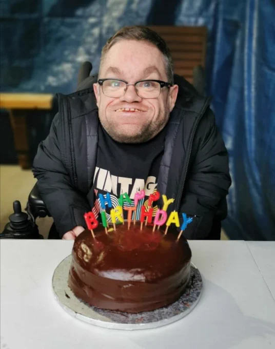 Dermot Devlin in front of a blue backdrop.

He is wearing a black coat and a t-shirt, which reads "Vintage 1979".

In front of him, is a big chocolate cake, with candles.

Each candle has a letter on it. Together, they read out "Happy Birthday"