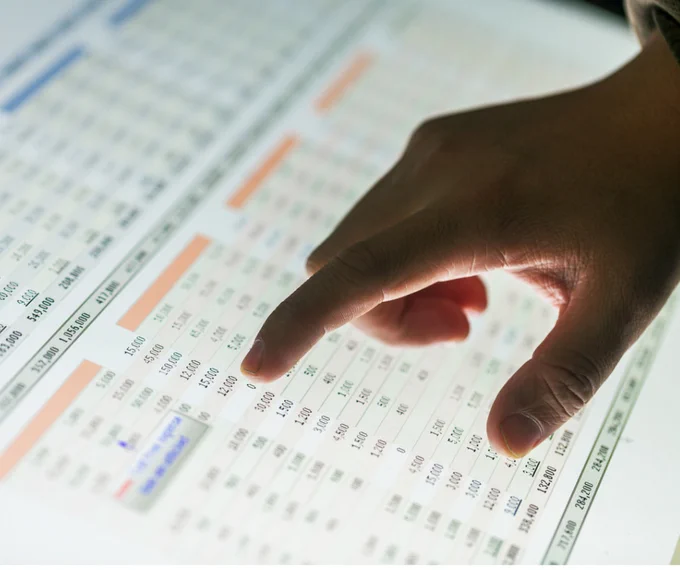 Hand points at spreadsheet on computer screen.