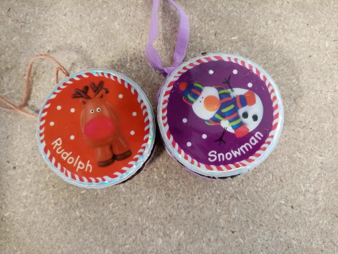A red Christmas decoration with Rudolph the red nosed reindeer on and a purple Christmas decoration with a snowman on.
