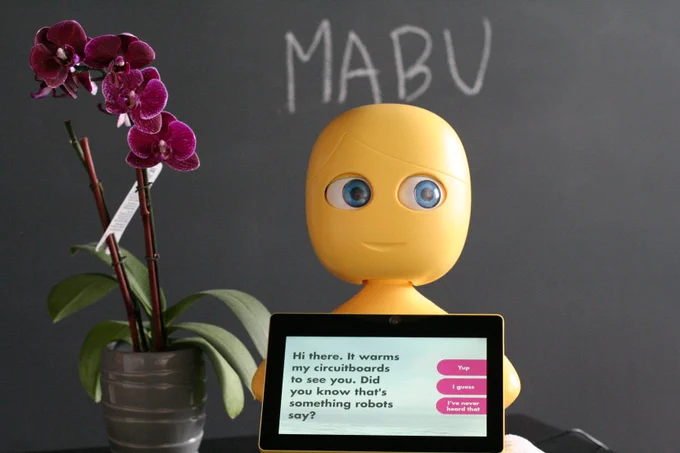 Mabu healthcare companion robot sitting in front of a blackboard with a message on her screen saying "Hi there. It warms my circuitboards to see you. Did you know that's something robots say?"
