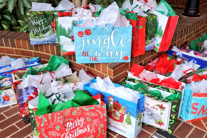A display of over a dozen large, string-handled Christmas gift bags of many designs, each filled with fluffy tissue paper in white, red, or green.