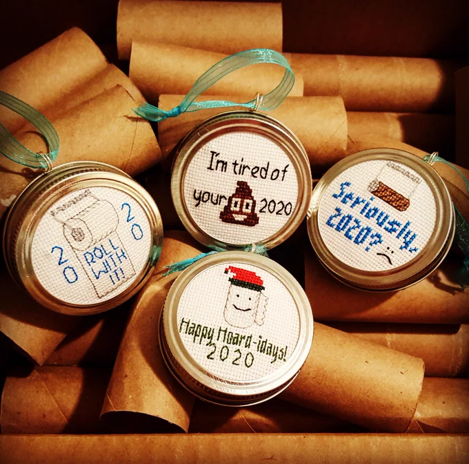 Several "Put a Lid on 2020" Ornaments from PinUp Preserves are displayed on a background of empty paper towel tubes. The cross-stitch ornaments are framed in actual canning lids and the designs include: an empty toilet paper roll with the text "Seriously, 2020?"; a full toilet paper roll with the text "ROLL WITH IT! 2020"; a roll of paper towels in a red and green cap with the text "Happy Hoard-idays! 2020"; and one with the text "I'm tired of your (poop emoji) 2020".