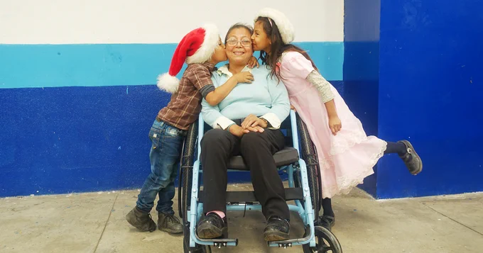 woman in wheelchair with young kids in santa hats kissing her on the cheek