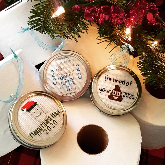 Three Put a Lid on 2020 Ornaments are displayed on rolls of toilet paper on a red and green plaid cloth. The ornaments are cross-stitch and framed in a silver canning lid with teal ribbon. L-R: a roll of paper towels in a red and green cap with the text "Happy Hoard-idays! 2020"; a roll of toilet paper on a silver holder with the text "ROLL WITH IT! 2020"; and "I'm tired of your (poop emoji) 2020". Branches of a Christmas tree with lights and garland peek out from the top right corner.