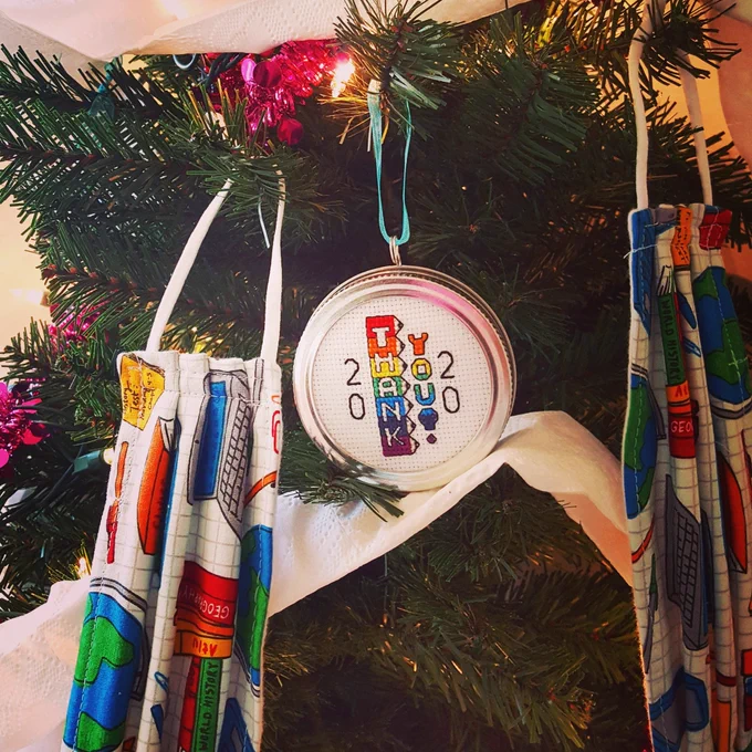 Two face masks hanging on a Christmas tree with a Put a Lid on 2020 Ornament in between. The mask fabrics has adorable brightly colored books and other school items displayed on a white graphing paper grid background, and the cross stitch ornament features stacked rainbow sharpened pencils that spell out "THANK YOU". The tree has white lights, pink tinsel garland, and white toilet paper garland.
