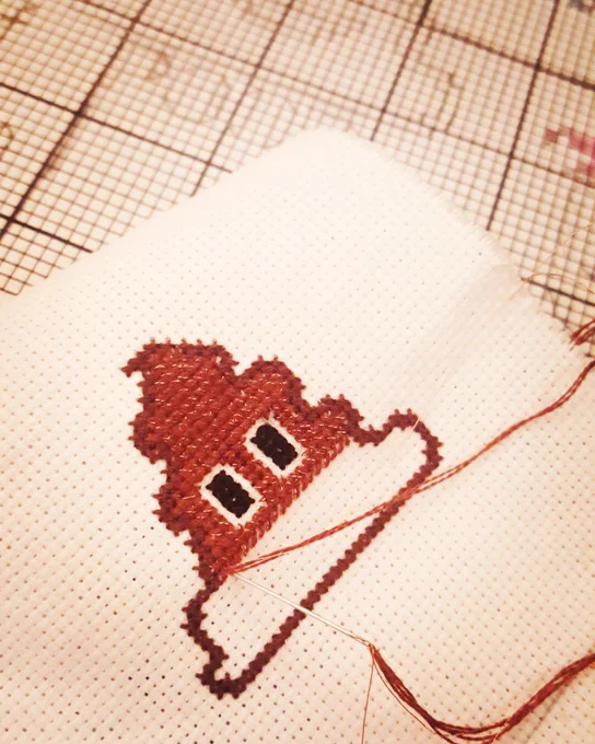 Close of of a cross stitch piece in progress. It is the poop emoji being sewn with sparkly brown thread, which is draped across the cloth mid-stitch. A grid paper is in the background with a pencil-drawn pattern.