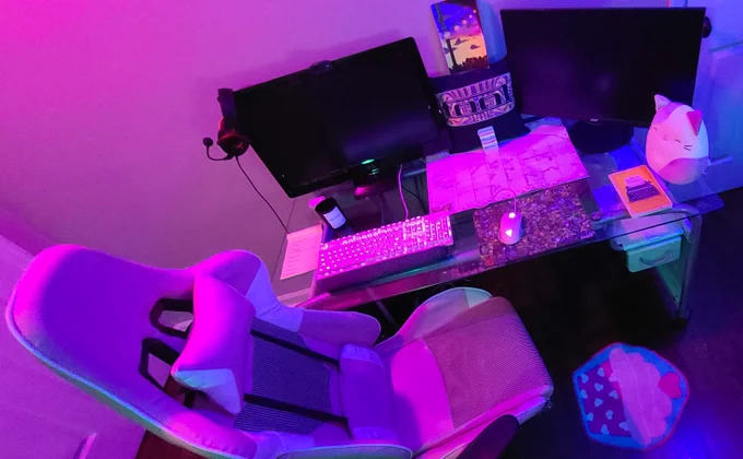 A desk with two monitors, a calendar, a stuffed animal cat, and a pillow. All of this is under a pink light.