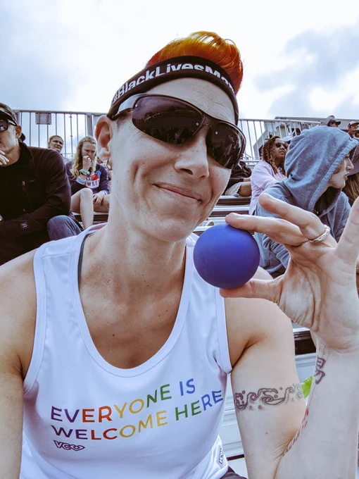 A cisgendered white woman sits on metal bleachers surrounded by people, wearing a white tank top that reads "Everyone is Welcome Here," a Black Lives Matter headband, sunglasses, smiling and holding a blue lacrosse ball