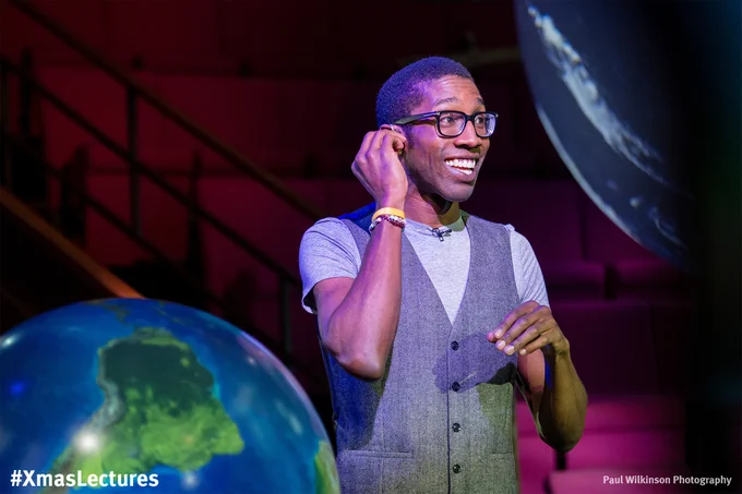 Man stood in an empty theatre between two globes of Earth, smiling and fitting an earpiece.