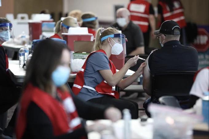 A blonde woman wearing a mask and face shield gives a COVID-19 vaccination shot to a man in all black wearing a hat. 