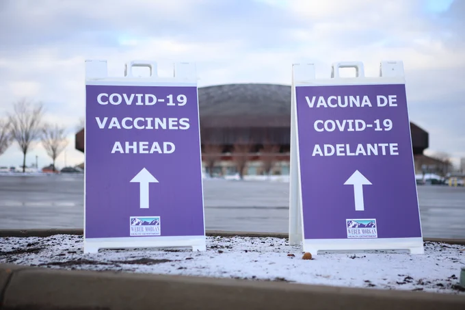 Two signs are pictured in the parking lot of the Dee Events Center. The sign on the left says "COVID-19 Vaccines Ahead" in English while the sign on the right says it in Spanish. 