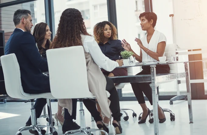 Company Team Members starting 2021 off with a relocation policy review. One man and four women sitting at a table, in business attire, discussing benchmarking their company's relocation policy to account for all of the changes relating to the COVID-19 pandemic.
