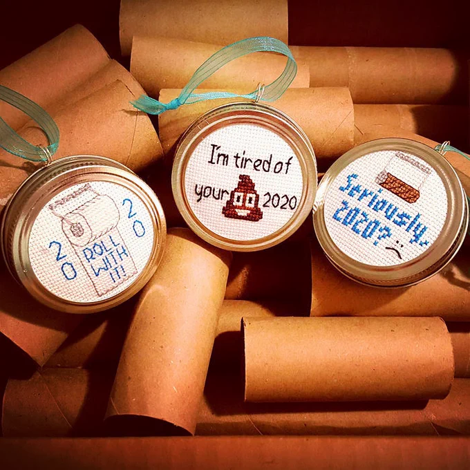 Several "Put a Lid on 2020" Ornaments from PinUp Preserves are displayed on a background of empty paper towel tubes. The cross-stitch ornaments are framed in actual canning lids and the designs include: an empty toilet paper roll with the text "Seriously, 2020?"; a full toilet paper roll with the text "ROLL WITH IT! 2020"; and one with the text "I'm tired of your (poop emoji) 2020".