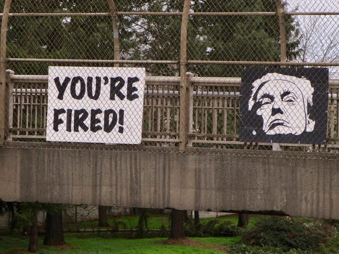 Painted cardboard signs hung above I-5 in Portland, Oregon.
One sign says "You're Fired!" and the other is a portrait of the failed IMPOTUS.