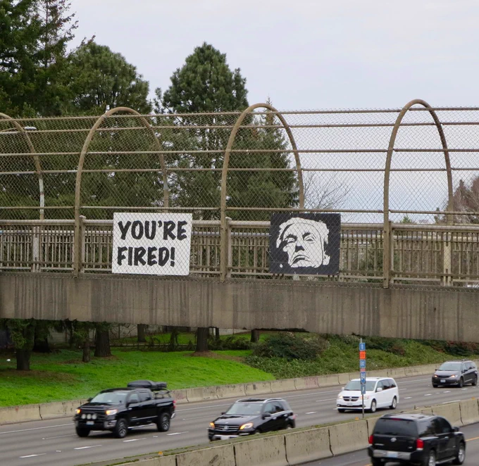 Painted cardboard signs hung above I-5 in Portland, Oregon.
One sign says "You're Fired!" and the other is a portrait of the failed IMPOTUS.