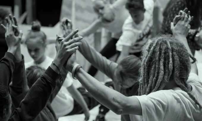 Black and white photo with a grey-blue filter: a black man with dreadlocks and a white t-shirt with his back to the camera stands in the foreground. He and a small group of people are holding hands, with their arms raised up in the air.