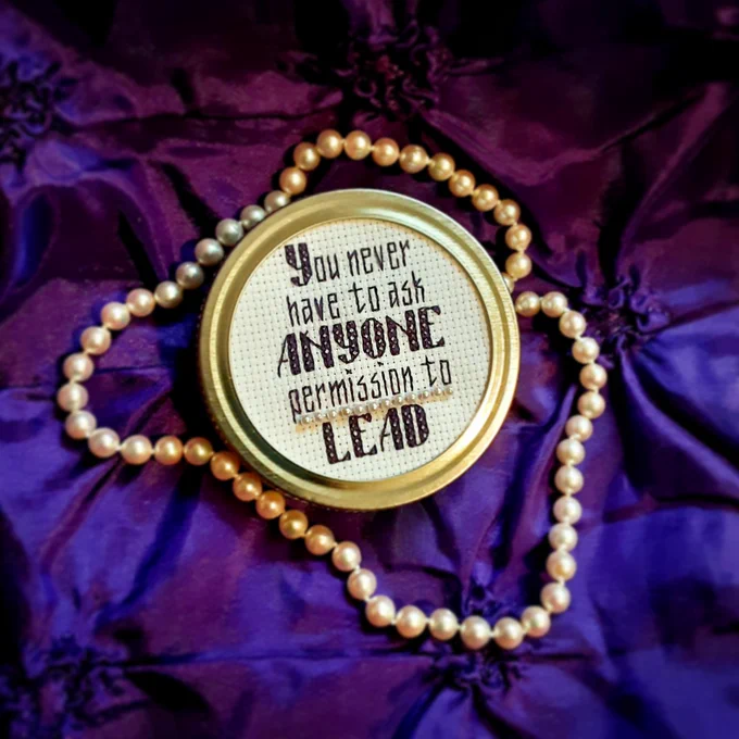 A small piece of cross stitch framed in a silver canning lid sits on dark purple fabric with a strand of pearls draped around it. Text: "You never have to ask ANYONE permission to LEAD". It is a quote from Vice President-elect Kamala Harris stitched in purple on white aida cloth, and "permission to" is underlined in pearls.