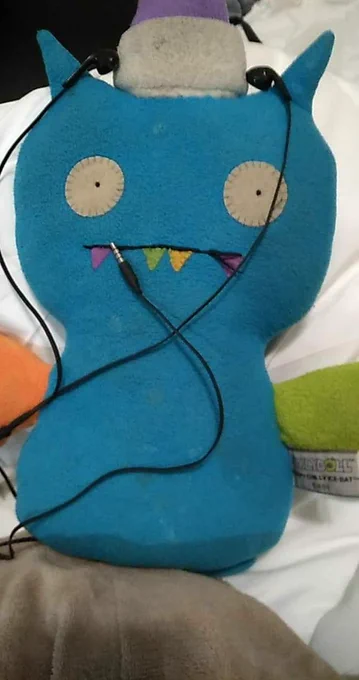 Photo of a blue felt stuffed animal bat with an orange wing, a green wing, and a purple hat. The hat has one eye and two teeth. The bat is wearing earbuds and the plug for them is by his mouth as if it's a microphone. He is laying on a white comforter.