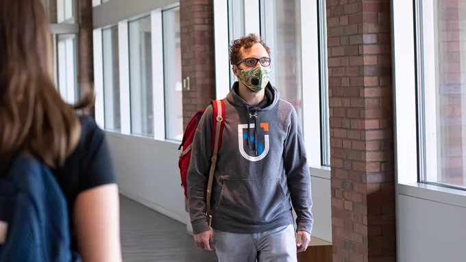 A male student wearing a grey hoodie with the university's logo walks down the hall wearing a light green mask with a kitten on it and a backpack slung over one shoulder.  