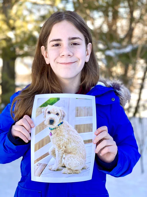 Gideon outside in the snow, holding a picture of a shaved down Soft-Coated Wheaten Terrier with ivory colored fur (Ollie). Ollie has short and curly hair. He is sitting at the top of a set of stairs on an outside deck. He has a green collar and a purple tag that says Ollie.