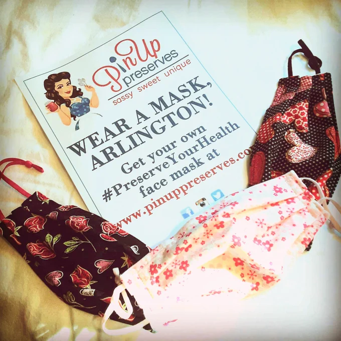 3 fabric face masks surround a paper saying "Wear a mask, Arlington! Get your own Preserve Your Health face mask at www.pinuporeserves.com"; the face masks are made from black fabric with red and pink roses and hearts, and white fabric with pink cherry blossoms.