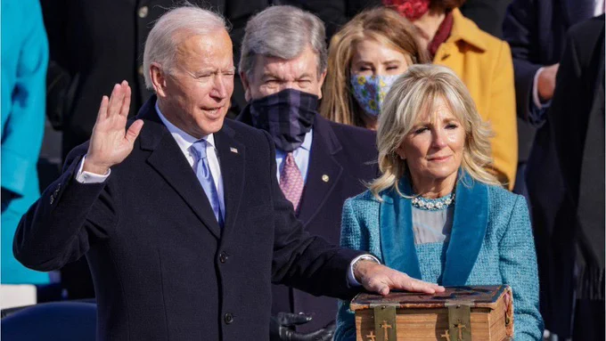 President Joe Biden a white man with white hair holds his hand on a bible and takes the oath of office. Jill Biden is next to him.