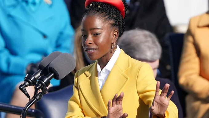 Amanda Gorman, a black woman wearing a yellow coat and a red headband reads a poem