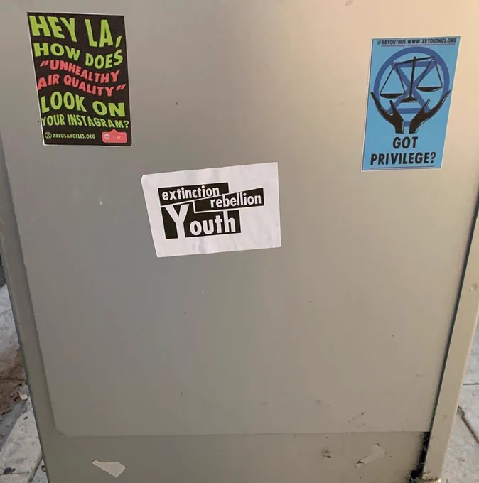 Three XRYLA stickers in a public area in Los Angeles. The sticker farthest to the left says “Hey LA how does “unhealthy air quality” look on your Instagram?  The sticker in the middle has the Extinction Rebellion logo on it. The sticker to the right is baby blue with the words “got privilege?”