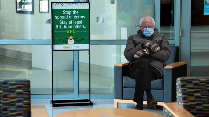 Seated Bernie Sanders with his mask and mittens. To his right, your left, is a sign stating "Stop the spread of germs. Stay at least 6ft. from others"