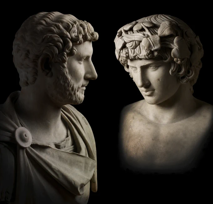 Two white marble busts showing the emperor Hadrian, who is bearded, facing to the right, and Antinous, facing to the left. The busts are against a black background.