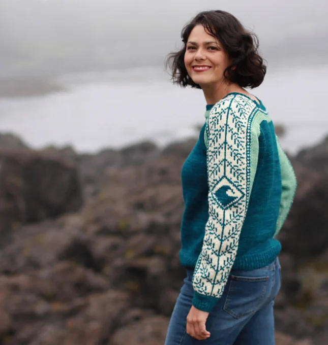 Model stands on a rocky shore wearing a pull over with colorblocks in the body and a panel of simple colorwork with pine branches and a wave down each sleeve. Sweater has ribbed cuffs and bottom and a boat neck.